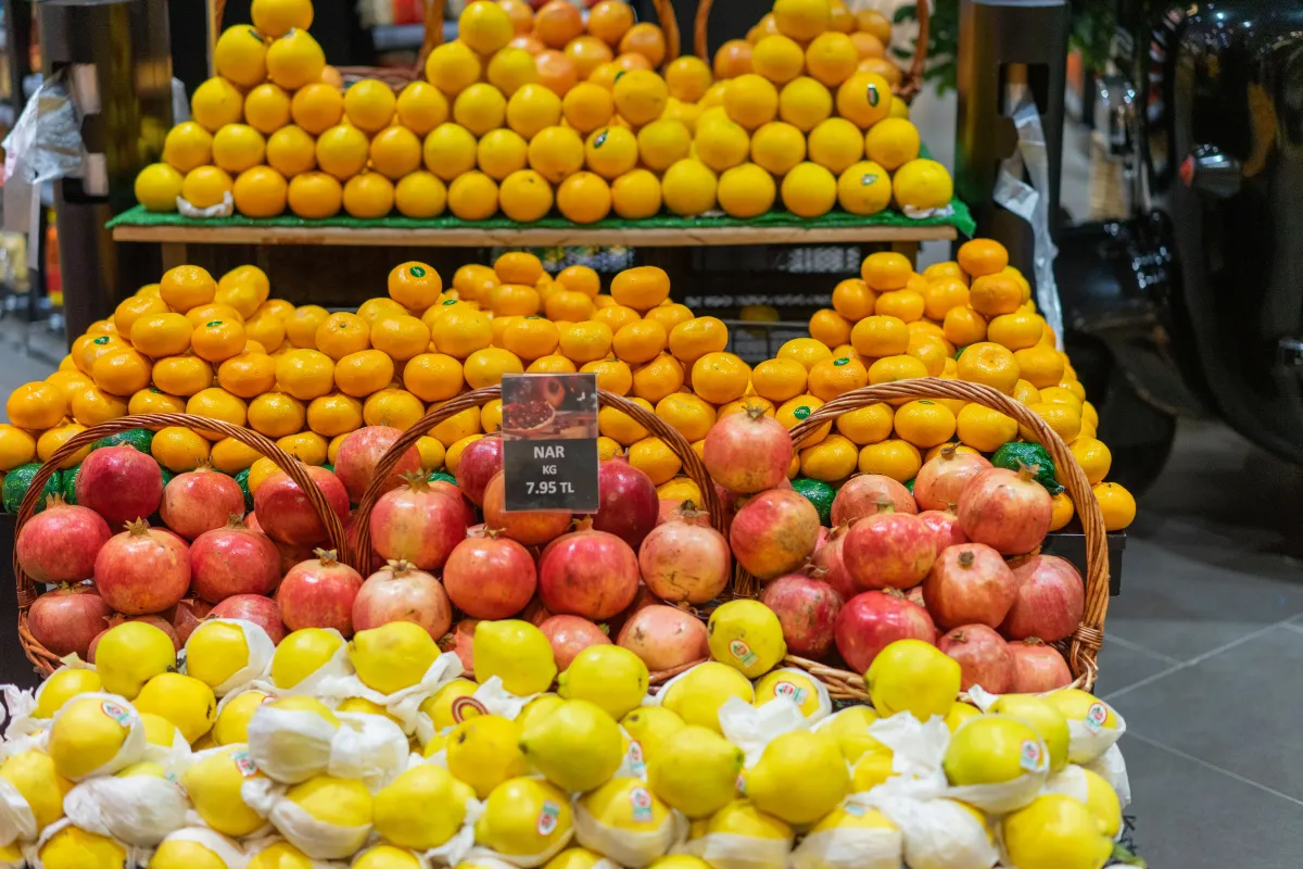 Bancada com diversas frutas em cestas, incluindo bananas, maçãs, uvas, peras e laranjas, em um mercado ao ar livre.