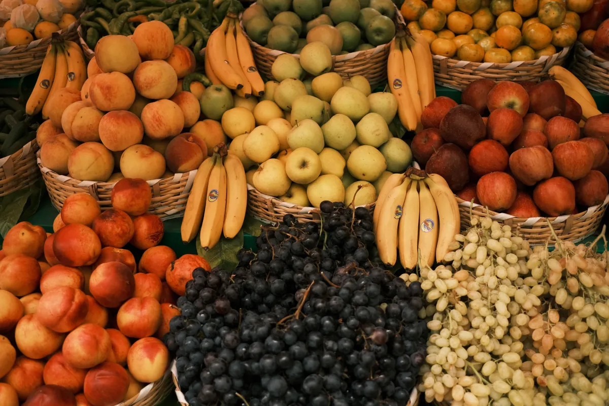 Bancas de frutas em mercado: cestas com maçãs e romãs ao lado de laranjas e outras frutas cítricas amarelas, com etiqueta de preço visível ao centro.