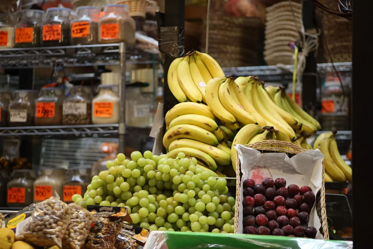 Cesta com bananas amarelas e cachos de uvas verdes e roxas em uma banca de mercado, com frutas frescas ao fundo em prateleiras de vidro.