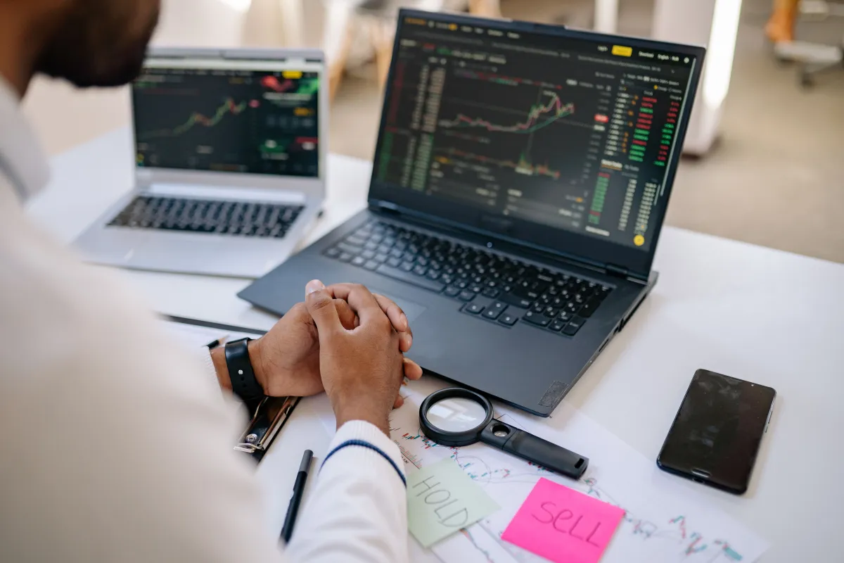 Pessoas trabalhando em computadores com gráficos e códigos na tela, enquanto analisam anotações e dados sobre investimentos em mesa de escritório, com celular ao lado.