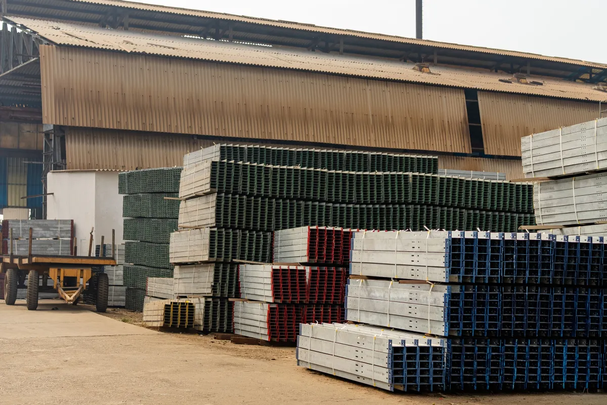 Pátio de armazém com empilhamento de caixas e blocos plásticos tipo grade, organizados em fileiras coloridas, com estrutura industrial ao fundo e carrinho de transporte na lateral.