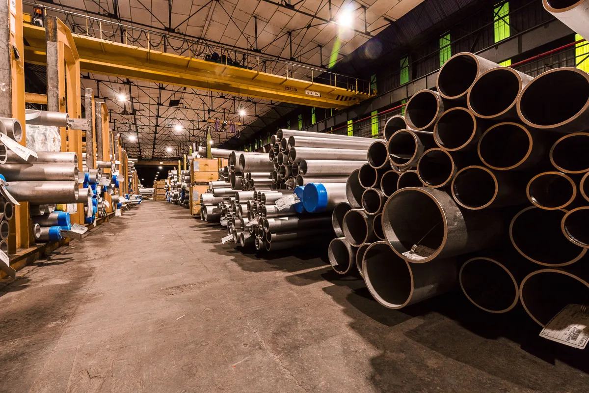 Armazém industrial com tubos de metal de diferentes diâmetros empilhados ao longo do corredor, estruturas de suporte e iluminação no teto, ideal para logística de materiais.