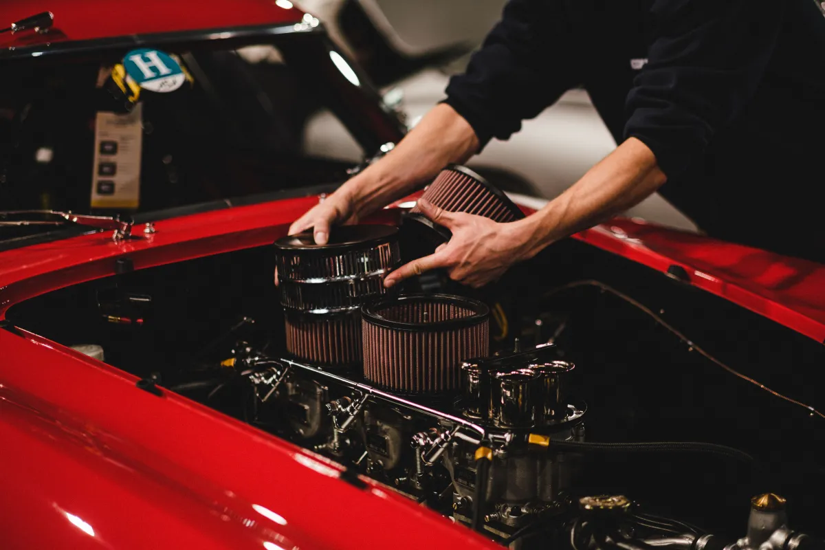 Mecânico instala filtros e componentes do motor em um carro vermelho, com as mãos trabalhando na parte superior do sistema mecânico no capô aberto, em oficina.