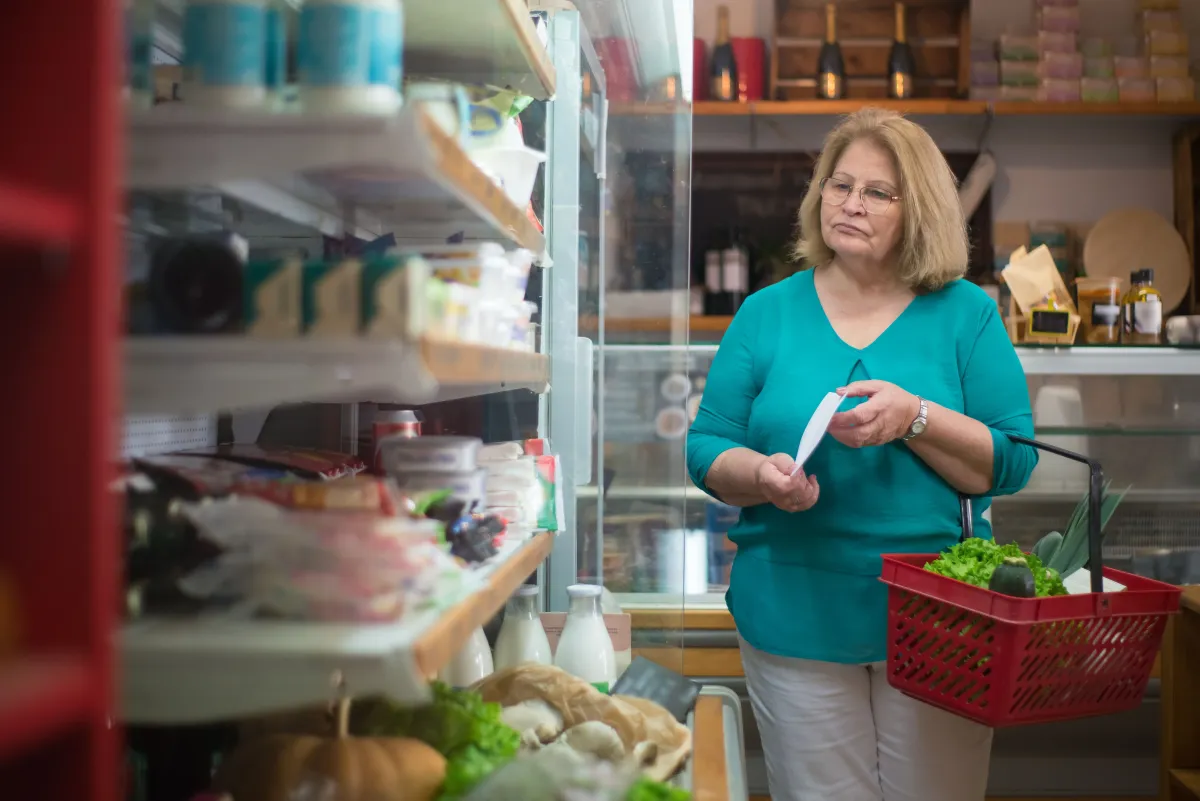 Mulher em uma mercearia ou mercado, segurando itens embalados e com uma cesta vermelha cheia de verduras, em frente à prateleira refrigerada