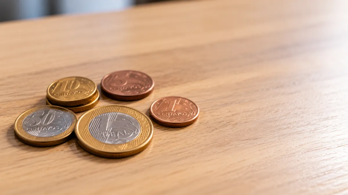 Moedas brasileiras de 1 real, 5 centavos, 10 centavos e 25 centavos sobre uma mesa de madeira, com close-up e luz natural suave.