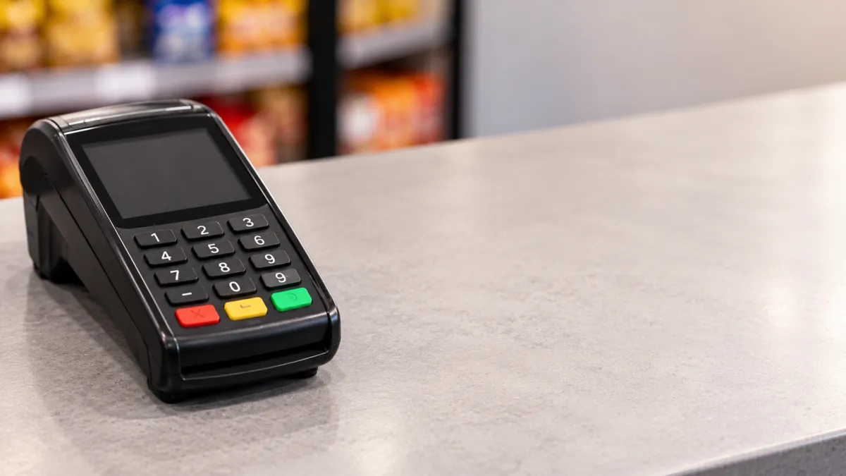 Terminal de pagamento com tela e teclado numérico sobre balcão, com botões de confirmação e cancelamento visíveis, em ambiente comercial desfocado ao fundo.