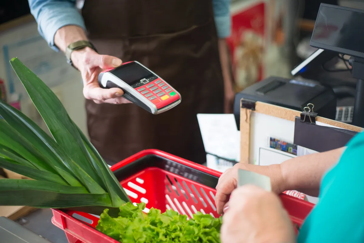 Pagamento com maquininha de cartão no caixa de mercado: cliente aproxima o cartão do leitor enquanto funcionários organizam as compras e há folhas verdes na frente