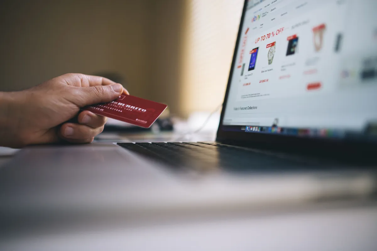Pessoa segura cartão de crédito vermelho em frente a um notebook com site de compras na tela, sugerindo pagamento online com segurança e transação digital.