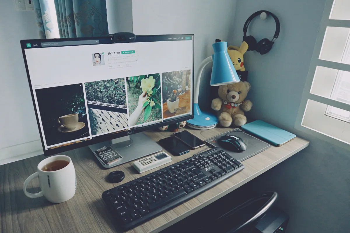 Mesa de trabalho com computador, teclado e mouse, tela exibindo layout de site, além de notebook, calculadora, caneca e acessórios decorativos como abajur e pelúcia em um ambiente