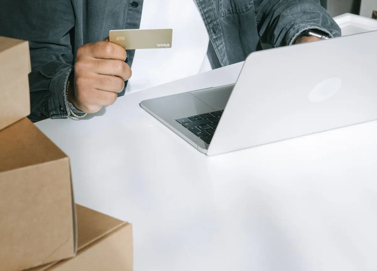 Pessoa em ambiente de compras online, usando notebook e segurando um cartão de crédito Visa para pagamento seguro, com caixas de envio ao lado.