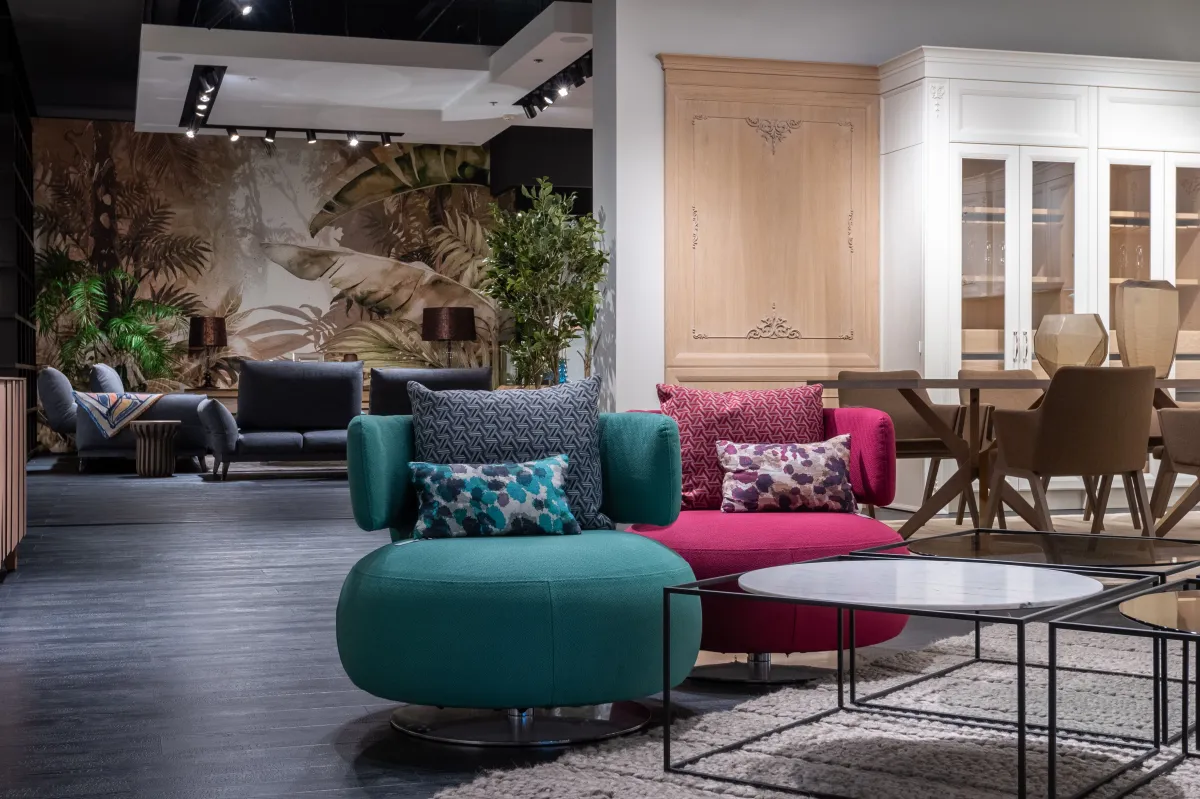 Sala de estar moderna com poltronas e sofás estofados, mesa de centro com tampo circular e tapete claro, ao fundo painel com arte de folhagens e móveis planejados