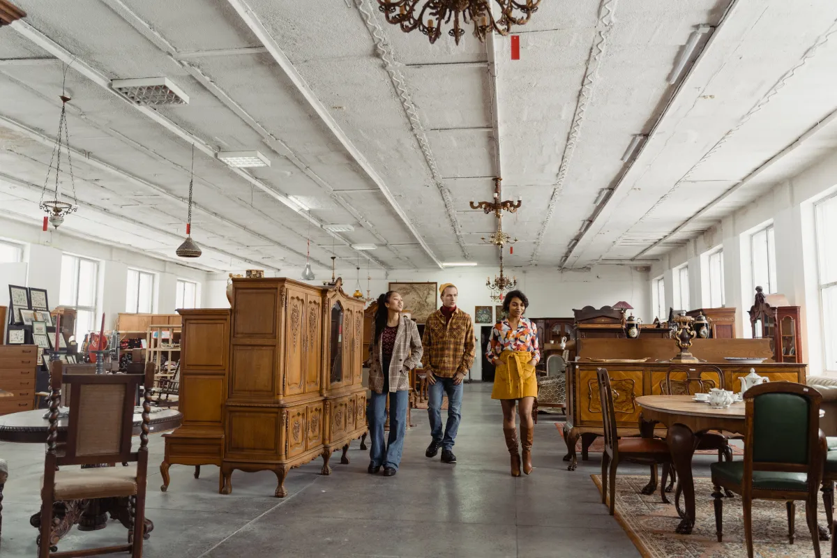 Interior de um salão amplo com móveis antigos de madeira, mesas e cadeiras clássicas, lustres e pessoas caminhando ao fundo entre vitrines e peças decorativas