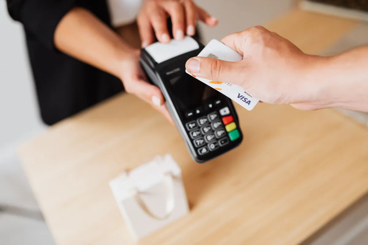 Pagamento com cartão de crédito Visa em um terminal POS: mãos de um cliente e de um atendente efetuam a compra, em ambiente interno.