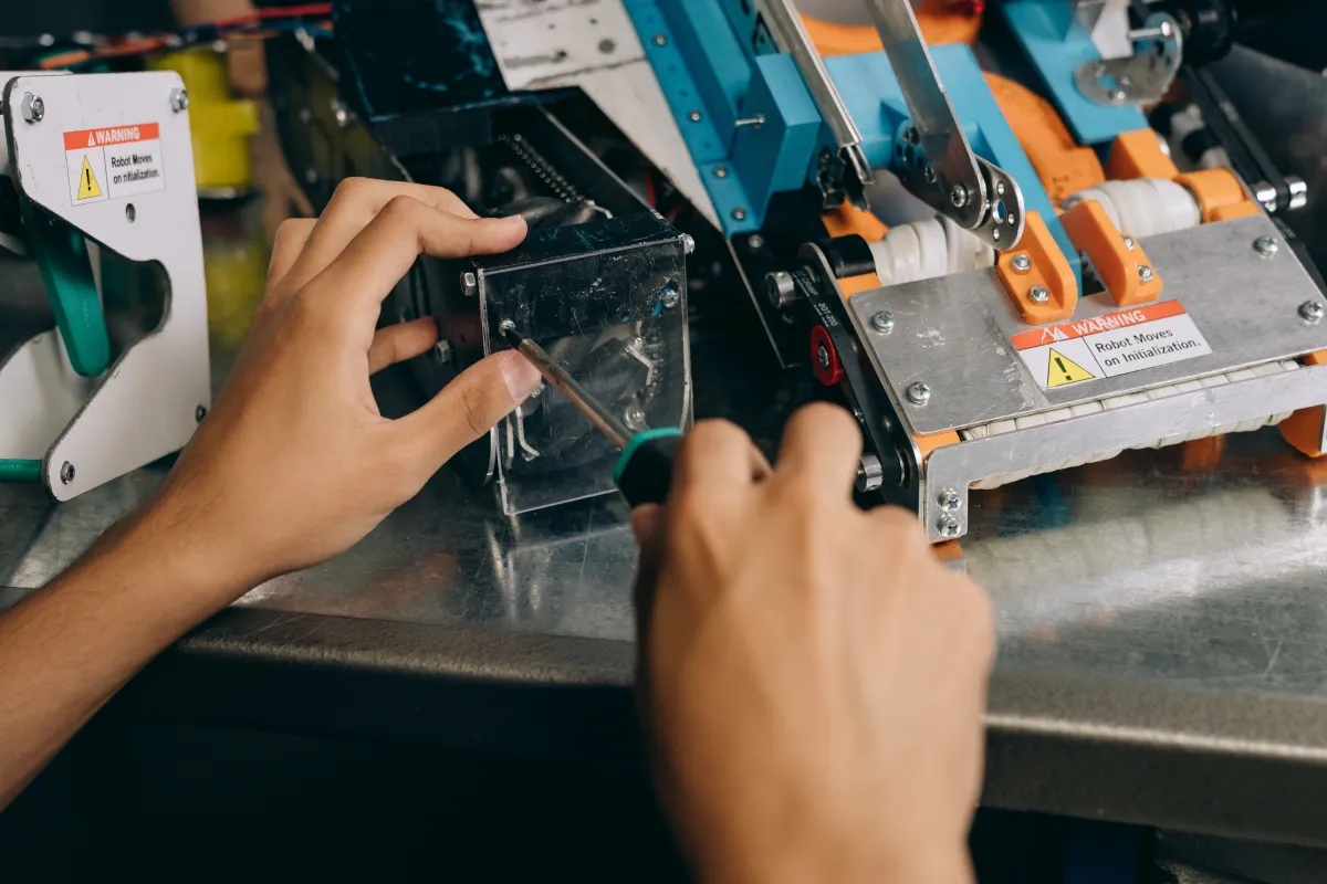 Pessoa ajustando uma peça em uma máquina industrial de automação, com mãos posicionadas em uma área de fixação e componentes metálicos ao redor.