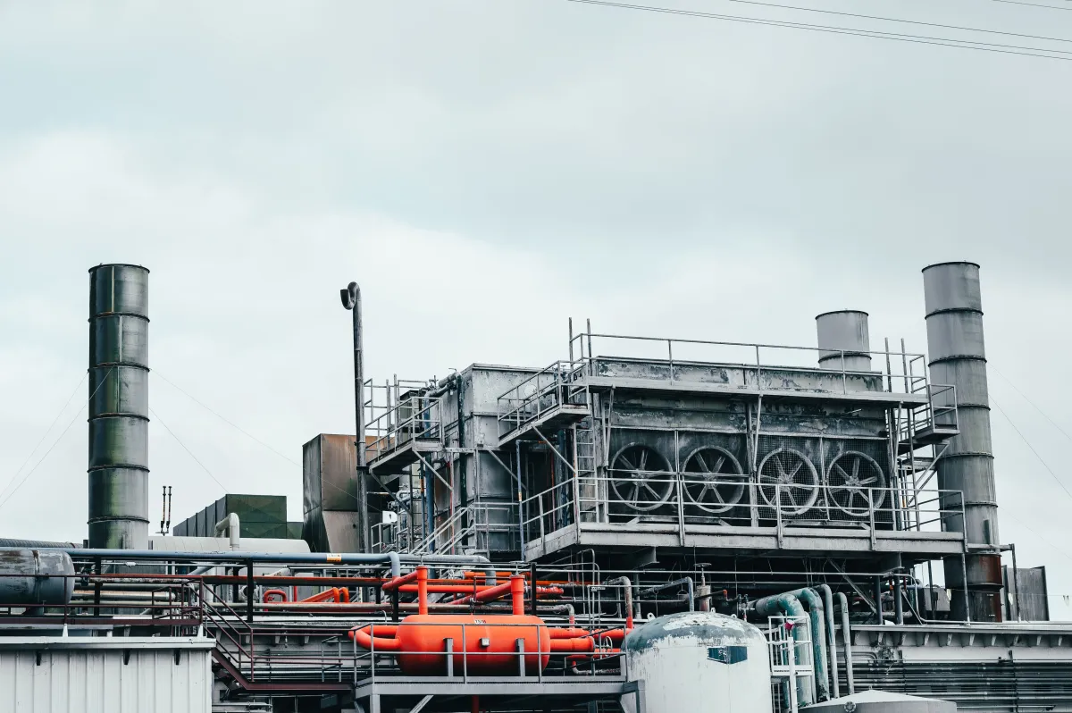 Vista de uma grande instalação industrial com chaminés de fumaça, tubulações e estruturas metálicas, com maquinário e equipamentos de processo em primeiro plano.