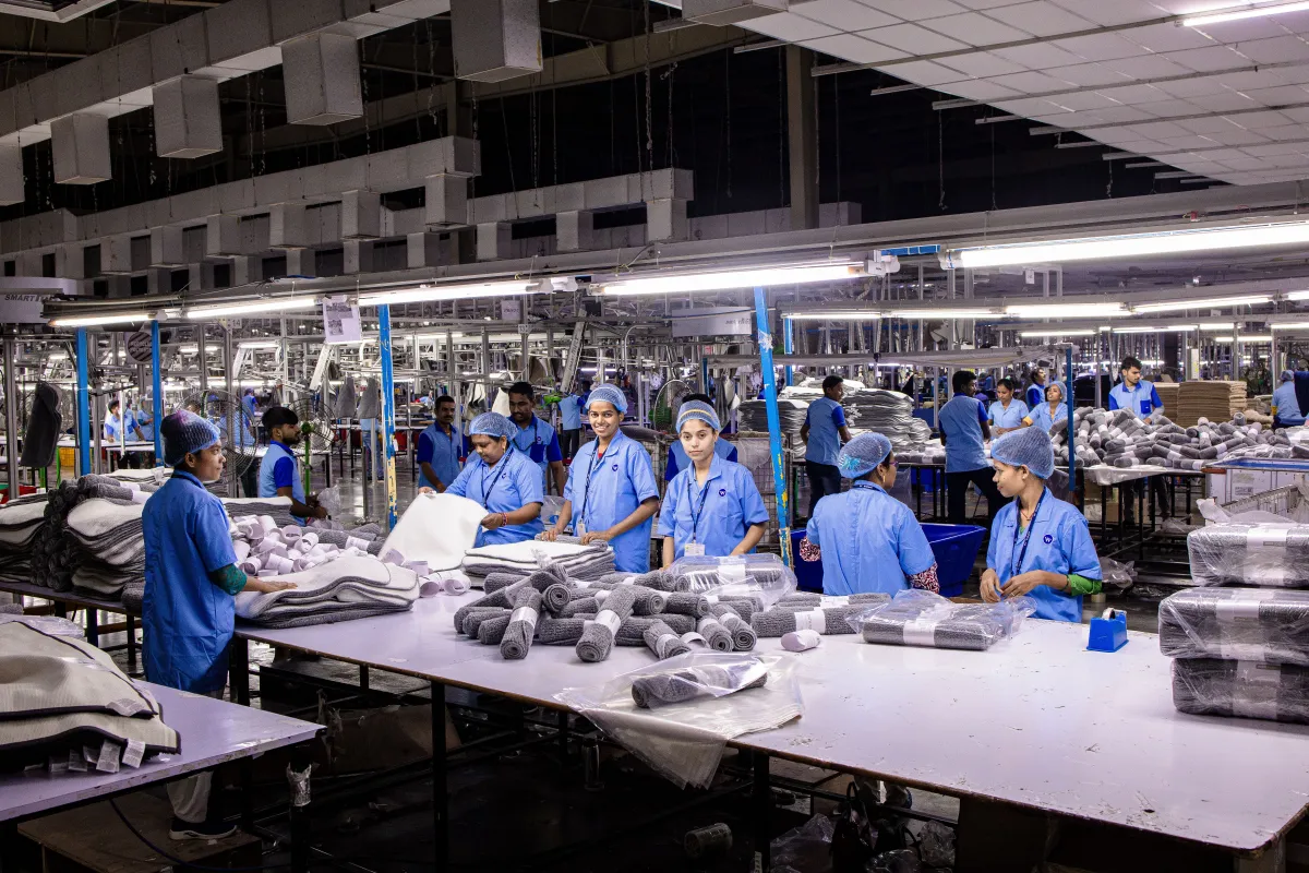 Equipe em linha de produção em fábrica, com trabalhadores usando uniforme e toucas, embalando componentes e organizando peças em bandejas e mesas de trabalho.