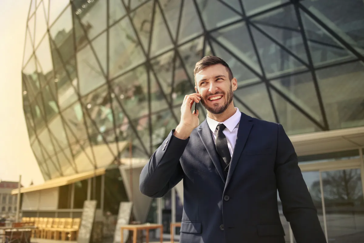 Homem de terno falando ao celular em frente a um prédio corporativo de vidro e metal, com sorriso ao ar livre no horário de dia