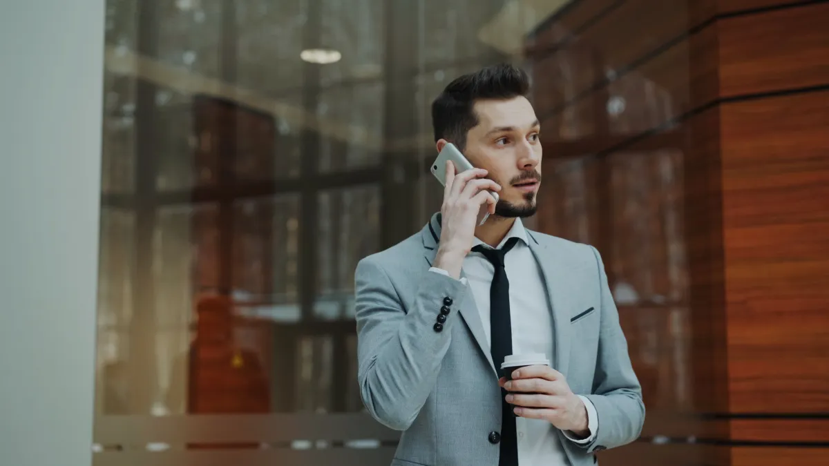 Homem usando smartphone em ambiente corporativo, segurando um copo com café enquanto fala ao telefone ao lado de uma fachada de vidro e madeira