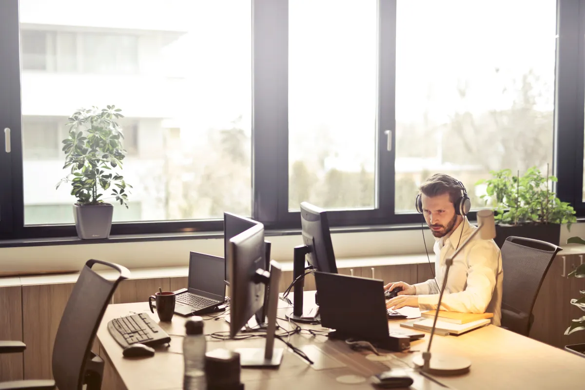 Homem trabalhando em um escritório moderno, com fones de ouvido, em frente a dois monitores e mesa organizada, aproveitando luz natural pela janela.