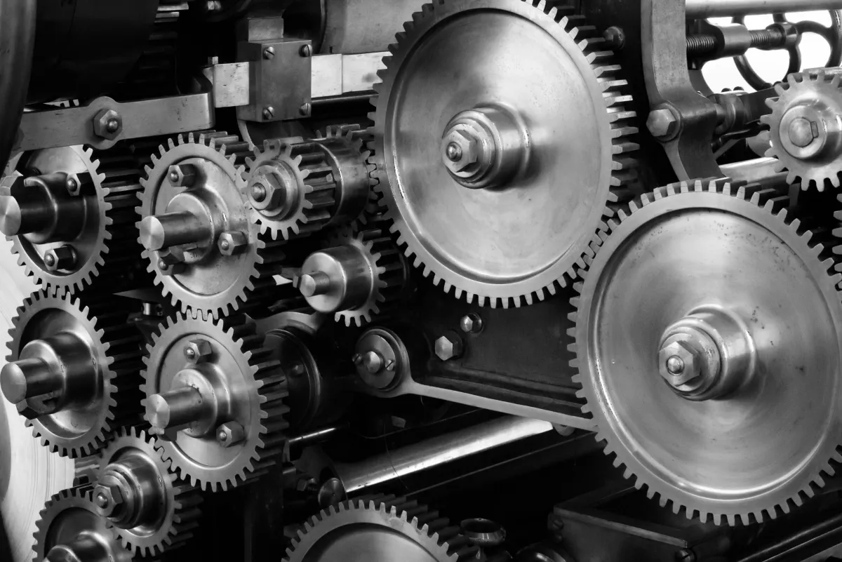 Imagem em preto e branco de engrenagens metálicas e rodas dentadas interligadas em uma máquina industrial, com parafusos e eixos visíveis.