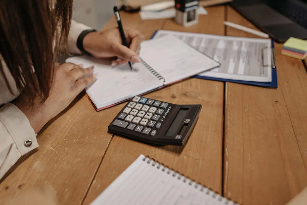 Pessoa escrevendo anotações em um caderno com calculadora digital sobre uma mesa de trabalho, ao lado de documentos e materiais de escritório, ambiente de escritório
