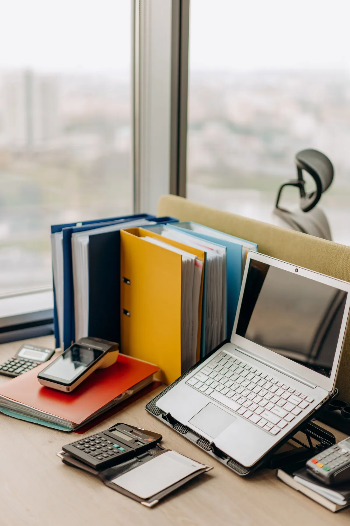 Mesa de escritório com notebook aberto, organizador de pastas coloridas, calculadora e cartões de acesso ao lado de documentos, em ambiente corporativo com janela ao fundo.