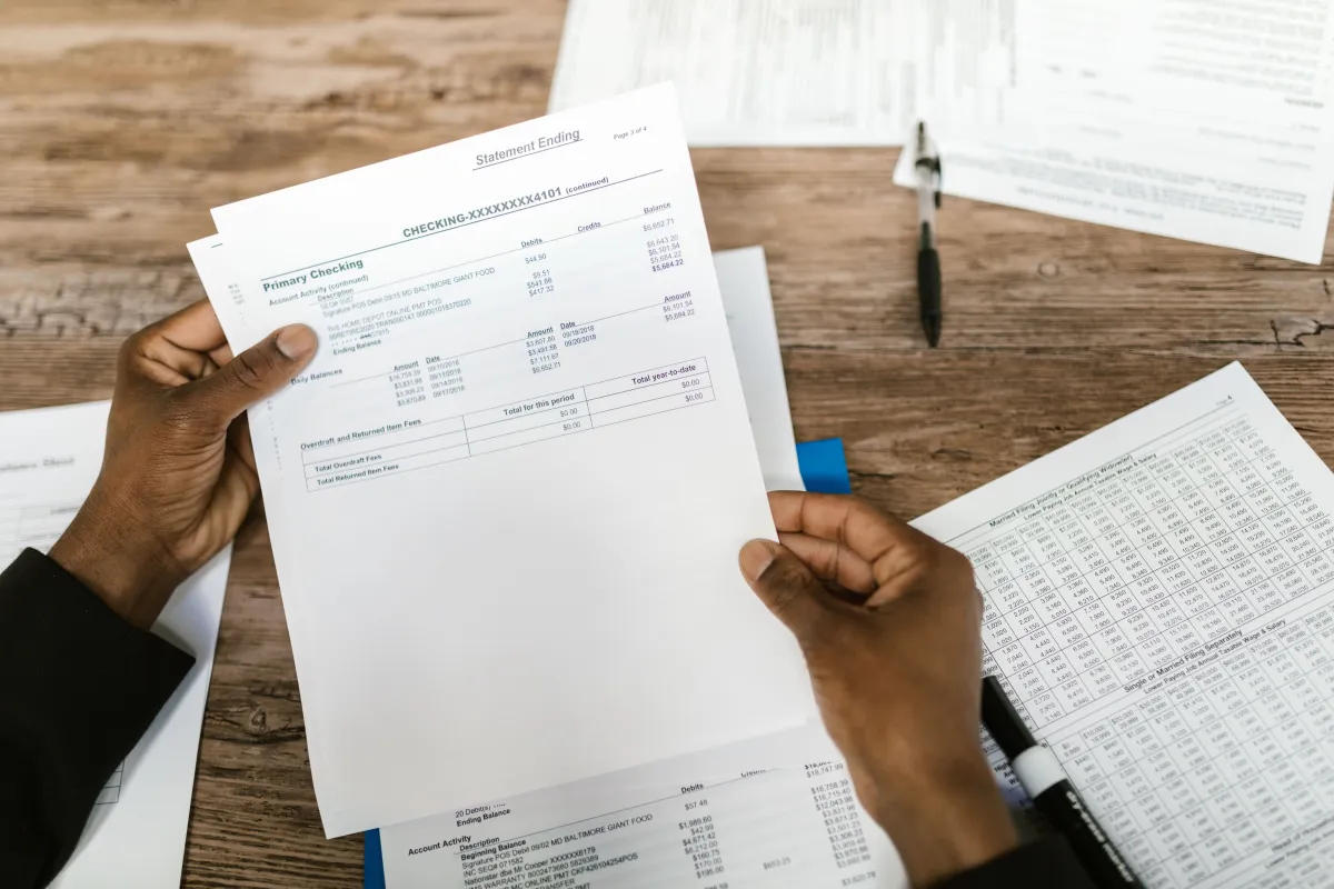 Mãos seguram documentos financeiros e formulários de checagem sobre uma mesa de madeira com caneta e planilhas ao fundo, sugerindo verificação e auditoria de dados.