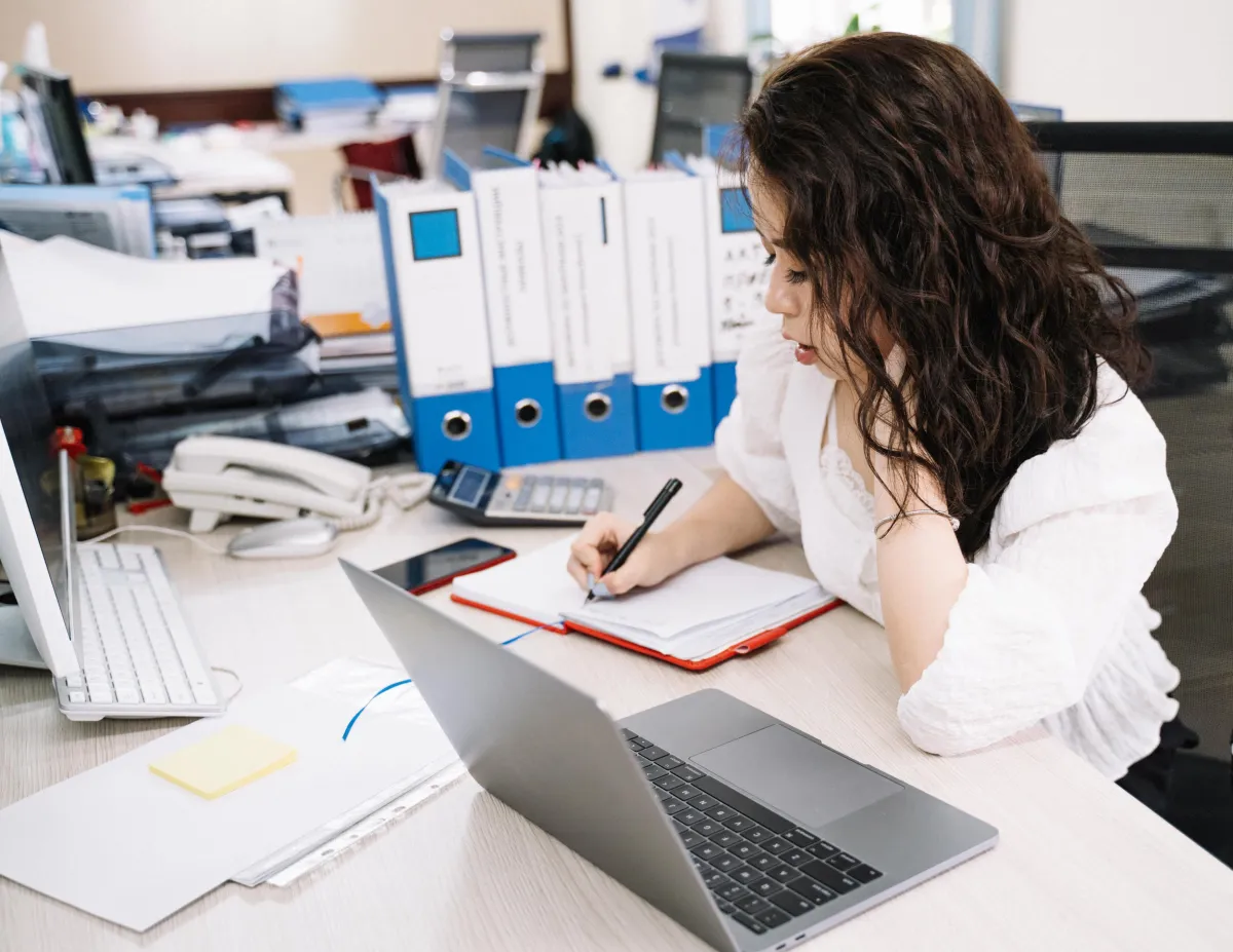 Mulher trabalhando em escritório corporativo, usando laptop e escrevendo em um bloco de anotações sobre mesa com pastas azuis e materiais organizados.