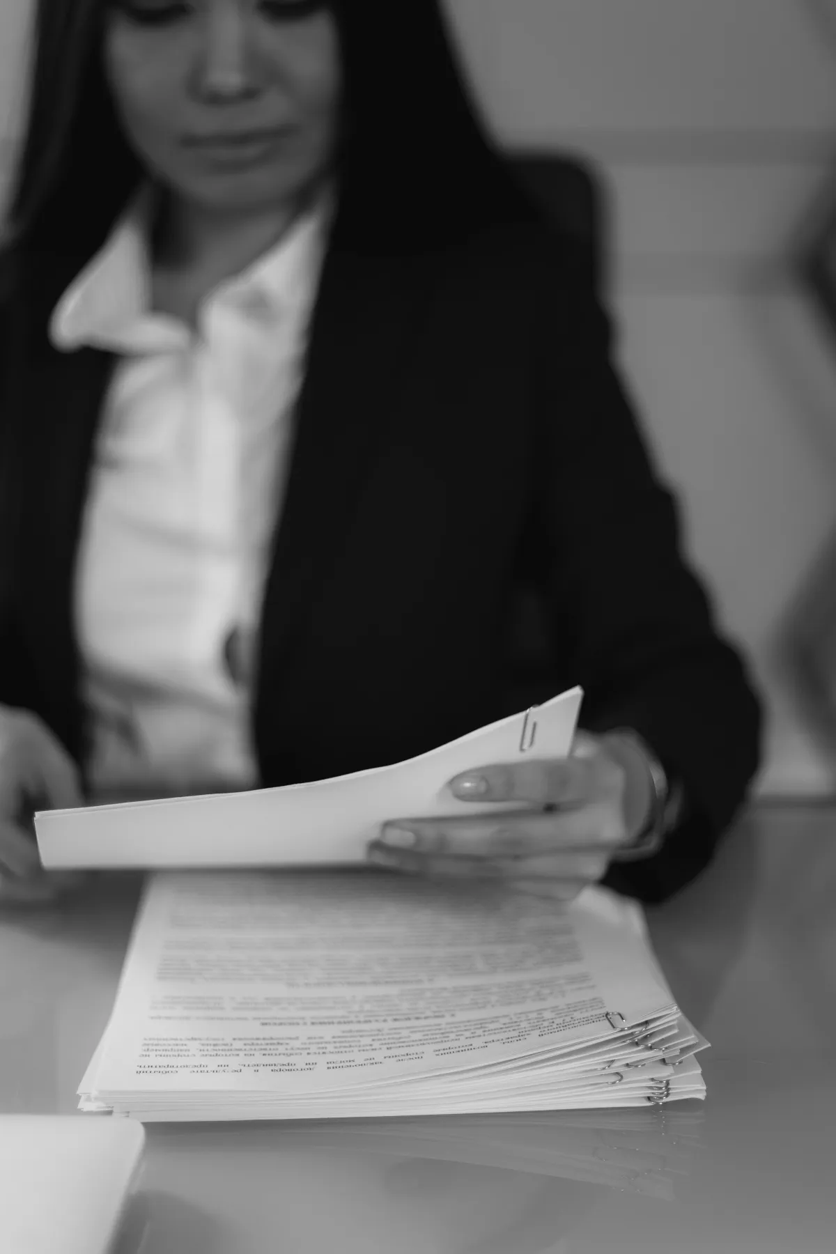 Pessoa em ambiente de escritório segurando documentos impressos e assinando ou revisando papéis, em imagem monocromática desfocada com livro de documentos na mesa.