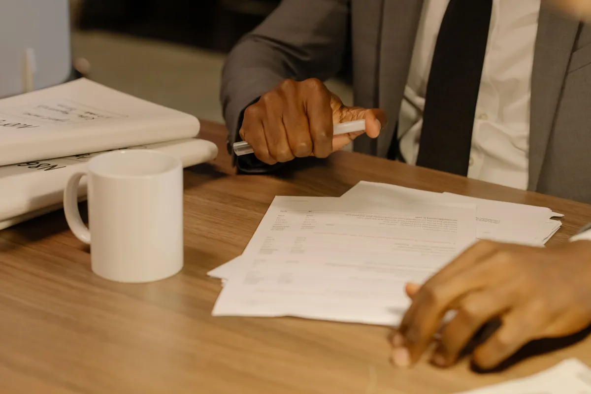 Mão de uma pessoa assinando documentos em uma mesa de trabalho com xícara branca e caneca ao lado, em ambiente interno com luz suave.