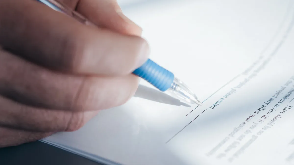 Pessoa escrevendo com caneta em um documento sobre uma mesa, com assinatura sendo feita em formulário e fundo desfocado ao redor.
