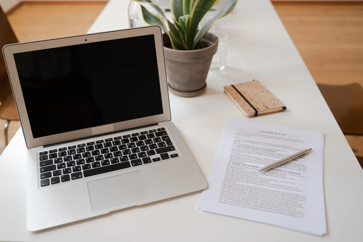Notebook aberto sobre uma mesa branca com teclado em destaque, ao lado de planta em vaso, bloco de anotações pequeno e documento impresso com caneta, em ambiente de trabalho.