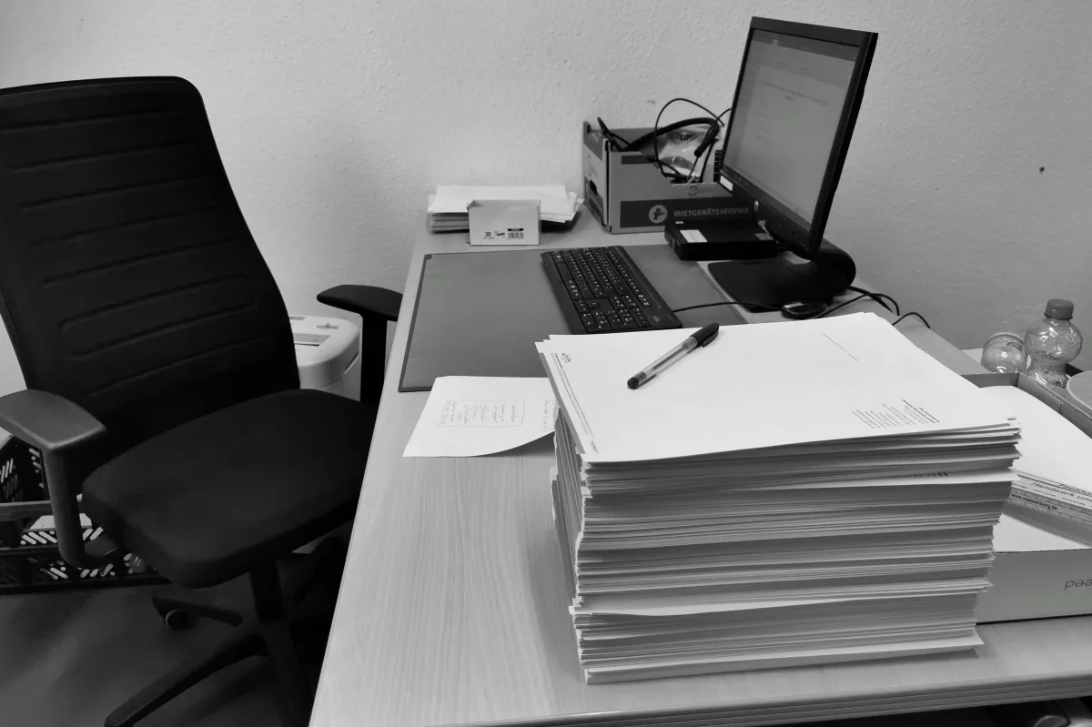 Escritório com mesa de trabalho organizada: pilhas de documentos e papéis, notebook ligado, teclado, caneta e itens de expediente em um ambiente de escritório simples e funcional.