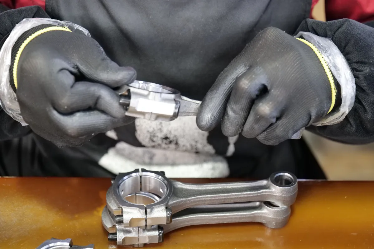Mecânico ajustando um motor com luvas de proteção, segurando uma peça metálica e utilizando uma ferramenta para montagem em bancada industrial.