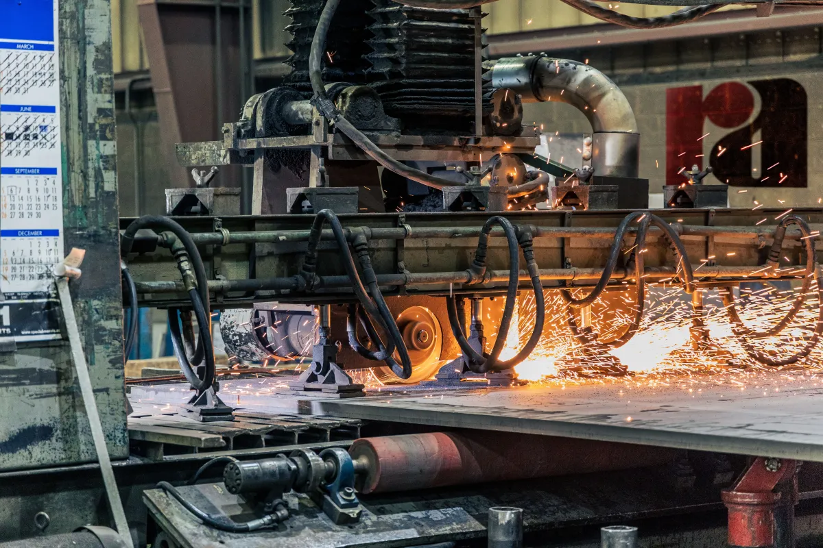 Detalhe de uma máquina industrial em uma oficina cortando ou processando metal, com faíscas e equipamento pesado em funcionamento.