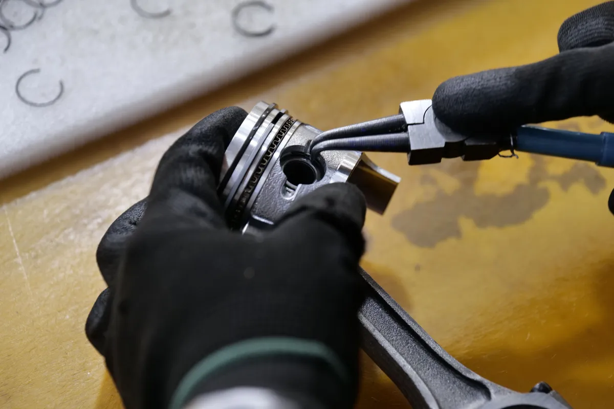 Montagem de um pistão e anéis em torno de peça metálica, com luvas de proteção e uso de ferramenta elétrica durante o reparo mecânico em bancada de trabalho.
