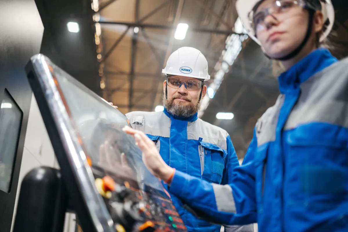 Trabalhadores de indústria com capacete branco e uniforme azul operam uma máquina em ambiente de fábrica, com foco no painel e no visor do equipamento.
