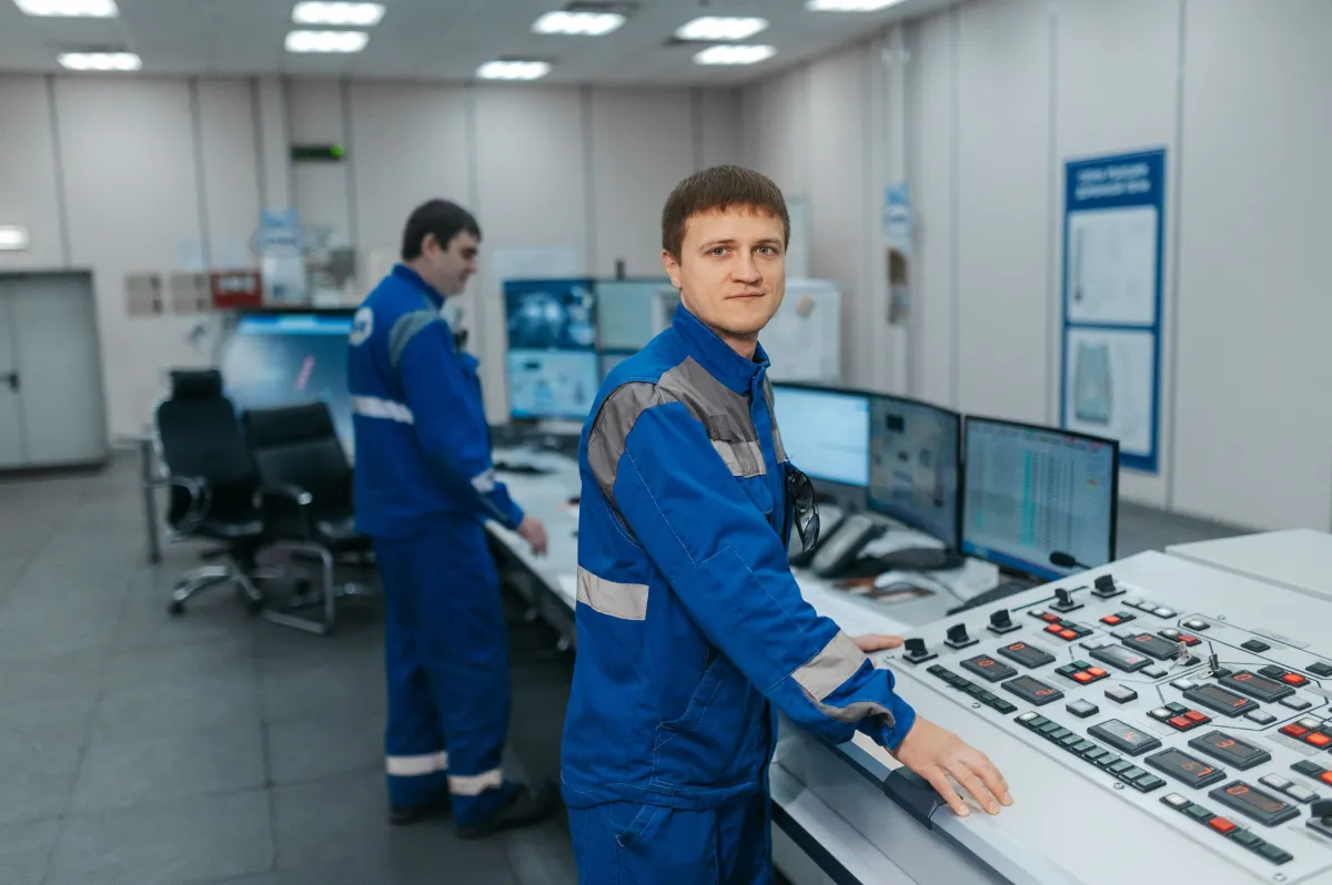 Operador de usina em sala de controle industrial, usando uniforme azul e trabalhando em um painel com botões, telas e sistemas de monitoramento ao fundo.