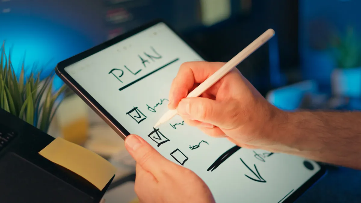 Pessoa segurando um tablet com desenho e anotações técnicas feitas com caneta stylus, em um ambiente de trabalho; gráficos e formas geométricas aparecem na tela em fundo branco.