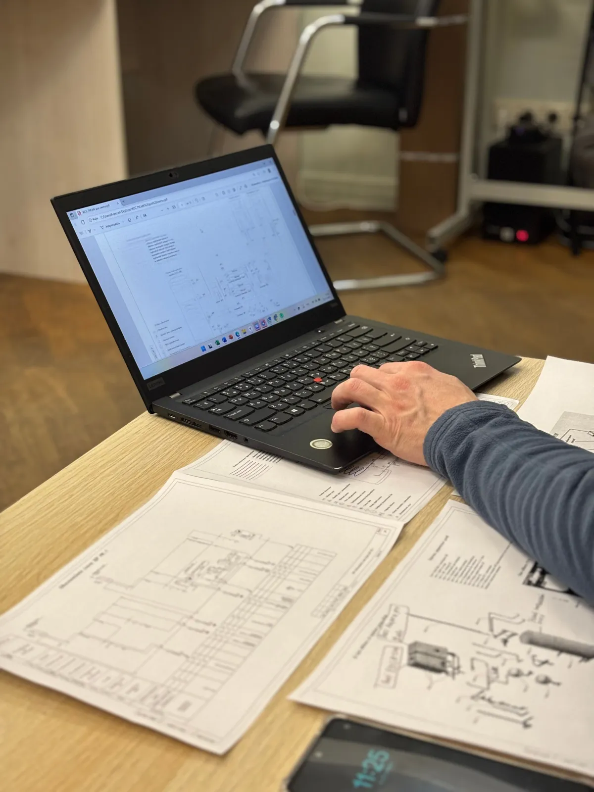 Pessoa trabalhando com notebook em uma mesa de escritório, com documentos técnicos e diagramas de engenharia abertos na tela enquanto a mão utiliza o mouse e há monitor ao fundo de