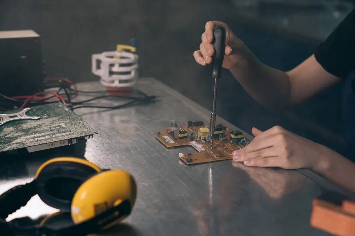Técnico utilizando uma chave de fenda para montar e ajustar uma placa de circuito eletrônico em uma bancada de eletrônica, com ferramentas e componentes ao lado.
