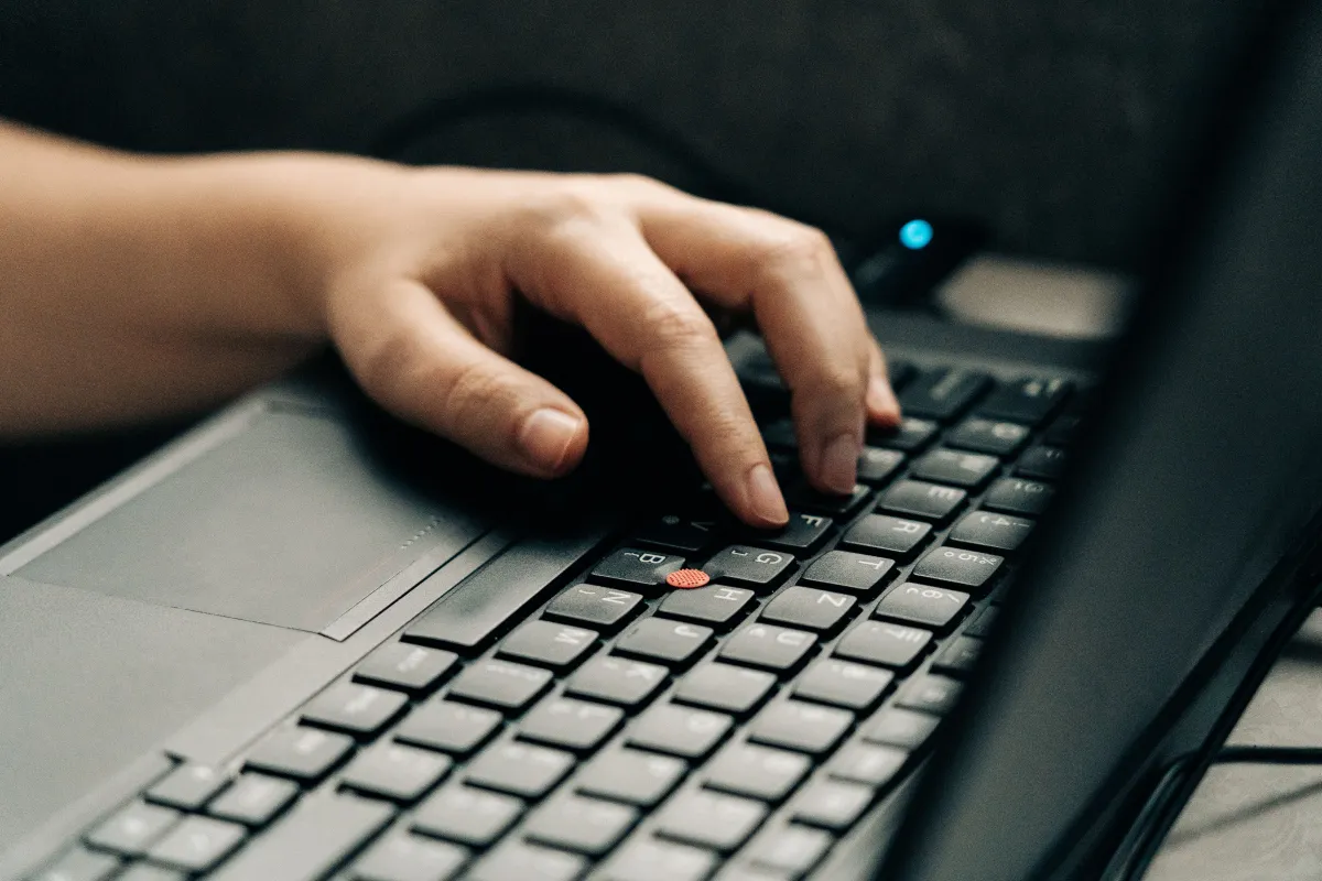 Mão digitando em um teclado de computador em ambiente de trabalho, com foco nos dedos pressionando as teclas e no dispositivo sobre uma mesa escura