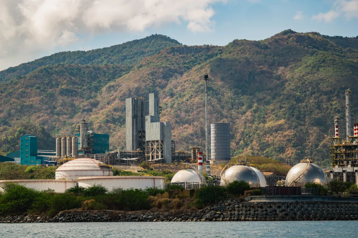 Vista de uma planta industrial com tanques e tubulações em uma usina à beira d’água, com montanhas ao fundo e estruturas metálicas no complexo