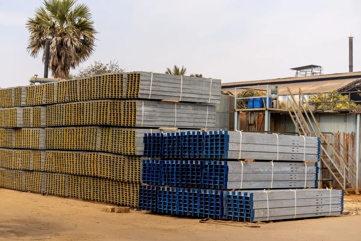 Pilhas de concreto pré-moldado e painéis de aço/estrutura empilhados ao ar livre em uma obra, com palmeiras ao fundo e área de trabalho com escadas e andaimes.