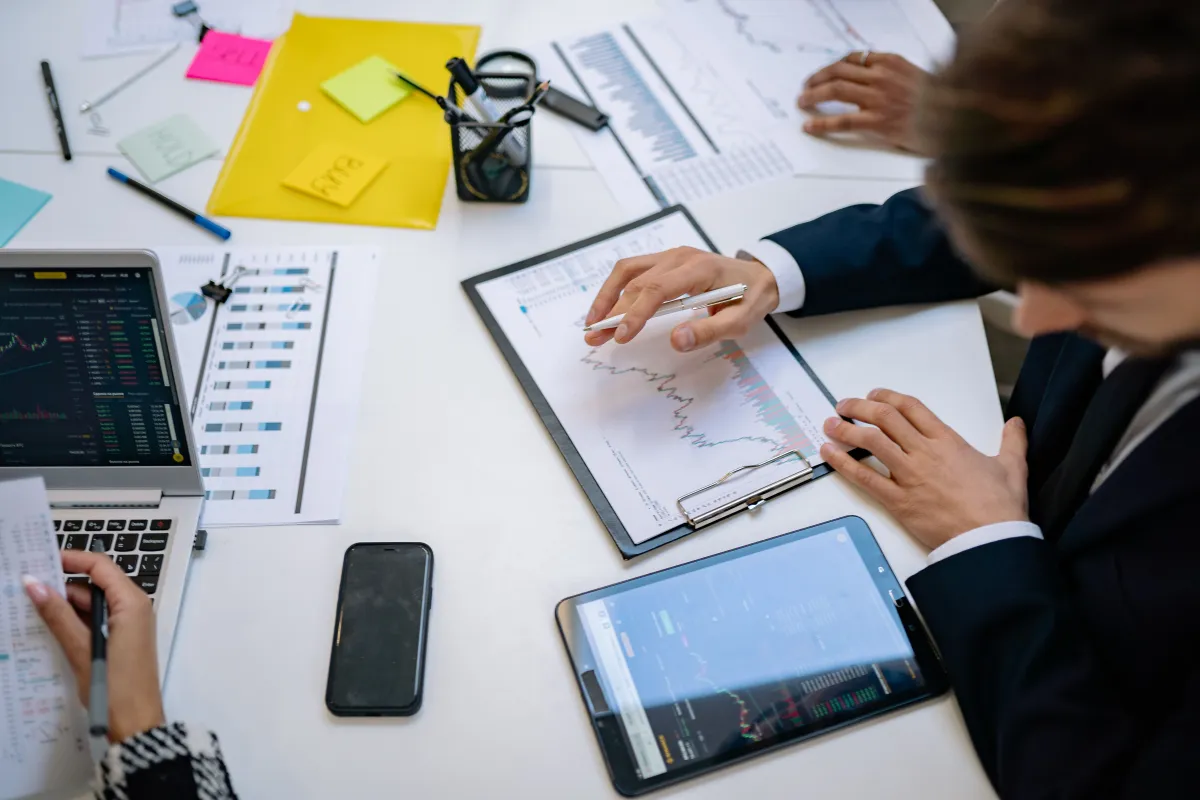 Equipe em reunião de negócios em uma mesa, com mãos analisando documentos e gráficos em tablets e computador portátil, mostrando organização e planejamento.
