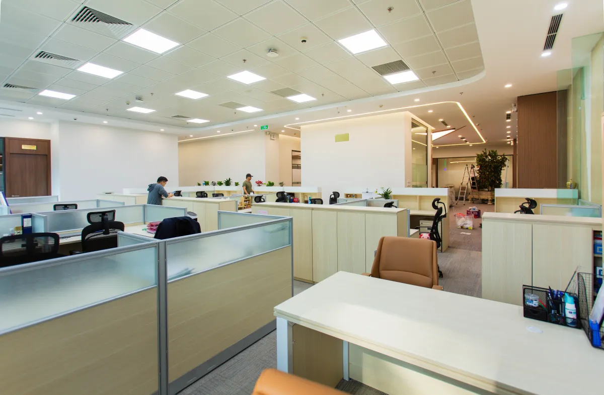 Ambiente de escritório corporativo moderno com baias de trabalho, divisórias de vidro, cadeiras ergonômicas e pessoas atendendo ao fundo, com iluminação no teto e mesas organizadas