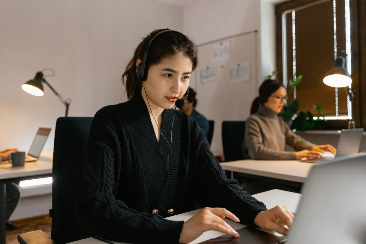 Mulher atendente em home office ou escritório, usando fone de ouvido com microfone e trabalhando no computador, com colegas ao fundo.