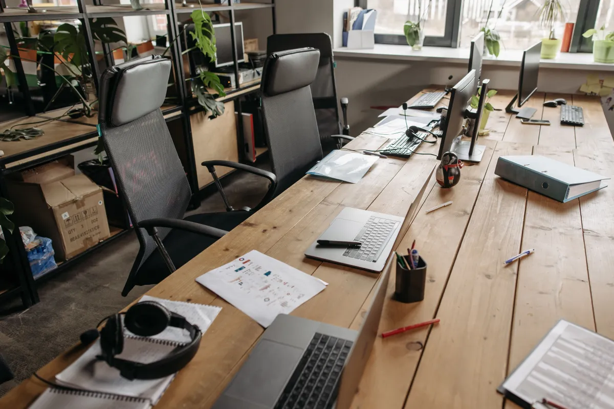 Ambiente de escritório com mesas de trabalho, computadores e papéis organizados, cadeiras giratórias e plantas decorativas em vasos perto das janelas, sala ampla e iluminada.