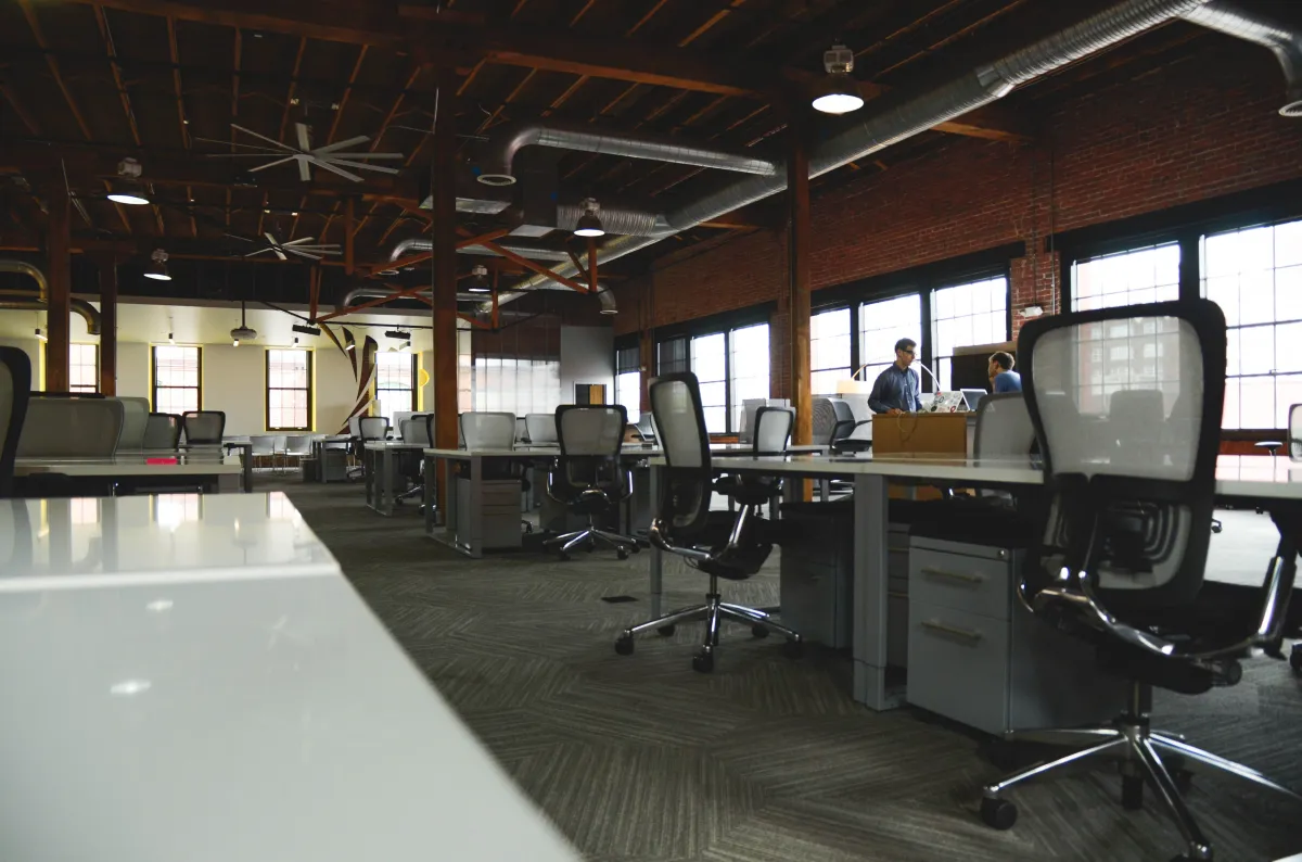 Ambiente de escritório corporativo amplo e moderno, com estações de trabalho, cadeiras ergonômicas, divisórias de vidro e iluminação industrial em teto com vigas metálicas.