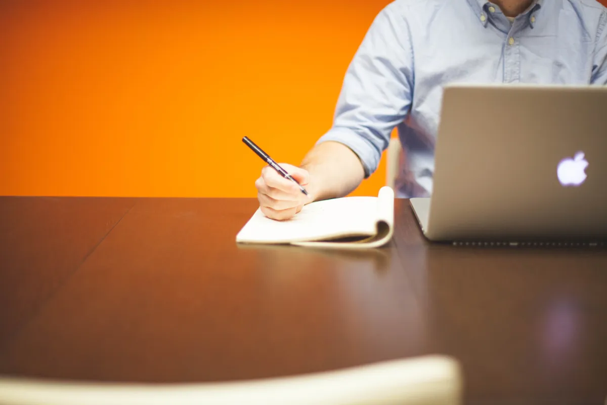 Pessoa trabalhando em escritório, escrevendo em um caderno e usando notebook em uma mesa com fundo laranja, destacando foco e produtividade no trabalho